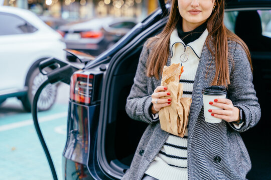People lifestyle and fast food concept. Young business woman sitting in a car trunk, waiting for charging.