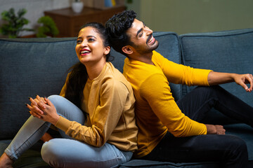Laughing indian couple talking each other by sitting back to back on sofa at home during evening -...