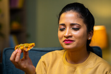 Young Indian woman enjoying byte of pizza at home during evening - concept of satisfied, foodie and...
