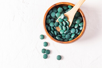 Round green spirulina algae pills in wooden bowl and spoon on white background with copy space. Top...