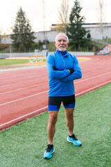 Obraz premium Fit elder athlete with gray beard in blue sportswear, ready for running on an overcast day at the track