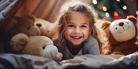 A charming cheerful little girl dressed in pajamas, playing with soft toys and bears on the bed, playing hide-and-seek, hiding under the blanket. The child is having fun at home. The concept of holida - obrazy, fototapety, plakaty