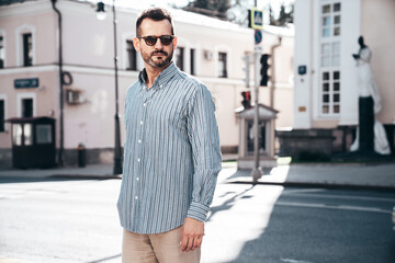 Portrait of handsome confident stylish hipster lambersexual model. Sexy modern man dressed in blue shirt and trousers. Fashion male posing on street background in Europe city at sunset. In sunglasses