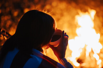 Backshot of a girl sitting near bonfire drinking warm tea or coffee. Spending time outdoors in...