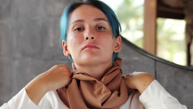 Portrait of woman in a hijab, she takes hold of headcovering with both hands and pulls it down with an effort, revealing her hair dyed blue. Looking straight to the camera aftermath