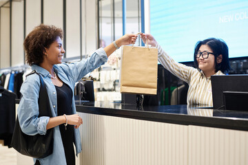 Happy african american woman taking bags with buys from smiling asian cashier at clothing store