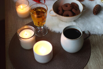 Cup of tea or coffee, glass of wine or juice, bowl of cookies, organic pomegranate, fluffy blanket, pine cones, various Christmas decorations and lit candles on the table. Christmas hygge concept.