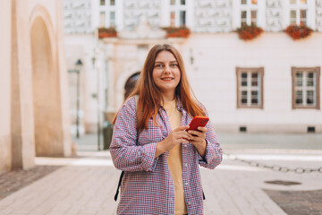 Naklejka premium Picture of pretty young woman staying on the street holding phone in hands. 30s tourist walking on old city street checks her smartphone. Use technology concept, Traveling Europe in summer