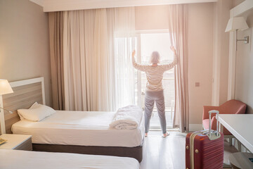 woman traveler opening the curtains and looking at the view from the window of a hotel room while...