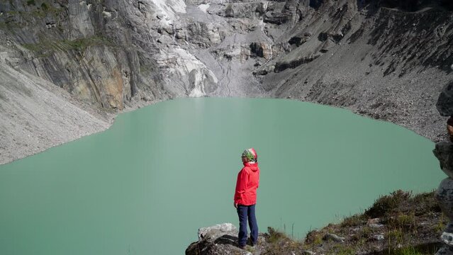 Female trekker dressed bright red jacket on the rock enjoying a glacier falling in high altitude Sabai Tso glacial lake cca 4350m. Makalu Barun National Park, Mera peak climbing route, Nepal. 4K video