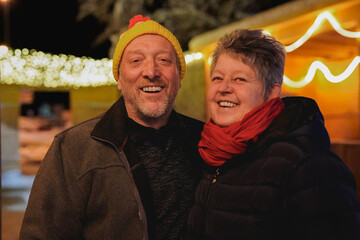 Happy senior couple smiling on camera at christmas market during winter time at night