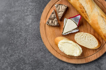 Processed triangle cheese in foil and baguette on wooden board on gray background.