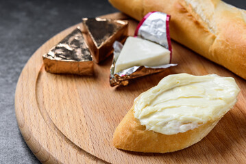 Processed triangle cheese in foil and baguette on wooden board on gray background.
