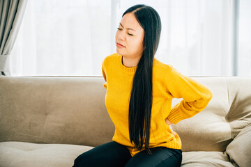 An Asian woman sitting on sofa holds her lower back in unbearable pain. Depicting chronic back pain issues discomfort and the need for medical care and attention. healthcare and problem concept - obrazy, fototapety, plakaty