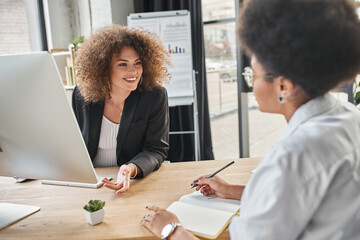 happy businesswoman talking to african american colleague with notebook in modern office, discussion