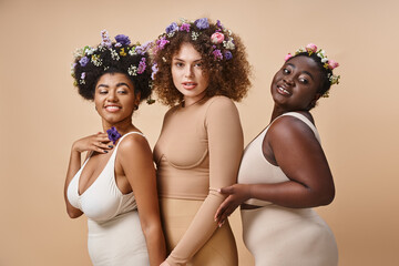 multiethnic plus size women in lingerie with colorful flowers in hair on beige, curvy elegance