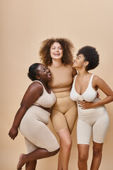 joyful multiracial women in underwear posing on beige backdrop, beauty and body positivity