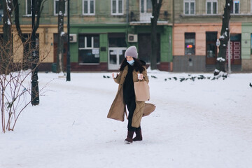 A young girl in a protective medical blue mask, a brown down jacket and a hat walks through a winter park with a bag in her hands and two glasses of coffee. Coronavirus Covid-19 pandemic. Ukraine Lviv