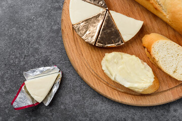 Processed triangle cheese in foil and baguette on wooden board on gray background.