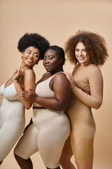 happy multiracial plus size girlfriends in lingerie smiling on beige backdrop, body positive beauty