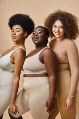 charming multiethnic plus size women in lingerie smiling at camera on beige backdrop, body positive