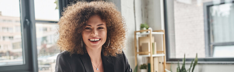 portrait of joyful businesswoman with curly hair looking at camera in modern office, banner - obrazy, fototapety, plakaty