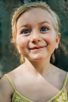 Close-up of a young girl in a yellow dress, smirking playfully with sparkling blue eyes, set against a blurred background