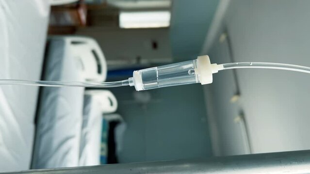 Drip chamber in an IV hanging on a stand against the background of blurred medical beds in a clinic ward. Vertical video