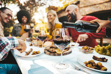 Happy friends having bbq dinner party in garden restaurant - Cheerful family drinking red wine together sitting at dining table - Food and beverage concept with guys and girls eating fresh meal - obrazy, fototapety, plakaty