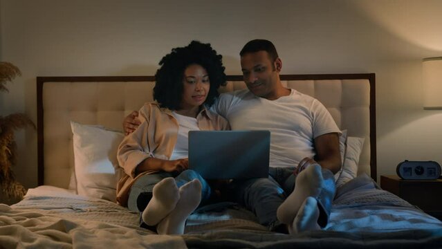Happy family African American married couple relax at evening at home bedroom on bed man and woman at night browsing laptop using computer talking discuss shopping together online watching movie film