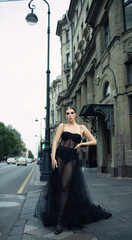A 30-year-old girl in a black transparent revealing dress on a city street. Style and elegance, femme fatale, photo shoot, advertising, girl in a black haute couture dress, close-up