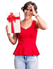 Beautiful young woman with short hair holding gift smiling happy doing ok sign with hand on eye looking through fingers