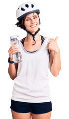 Beautiful young woman with short hair wearing bike helmet and holding water bottle smiling happy and positive, thumb up doing excellent and approval sign