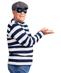Senior handsome man wearing burglar mask and t-shirt pointing aside with hands open palms showing copy space, presenting advertisement smiling excited happy
