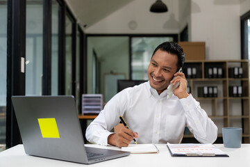 Obraz premium Handsome and happy Asian businessman using his mobile phone at the table Using mobile phone looking at smartphone working in company office with laptop computer technology