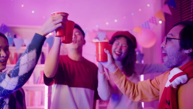 Friends Sit At Sofa Couch And Holding Drinks Joy Night Party With Pink Lighting At House Party.Young Asian Friends In Smart Casualwear Raising Arms While Dancing Together At Home Party In Living-room.