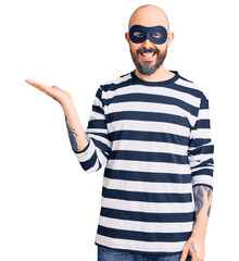 Naklejka premium Young handsome man wearing burglar mask smiling cheerful presenting and pointing with palm of hand looking at the camera.