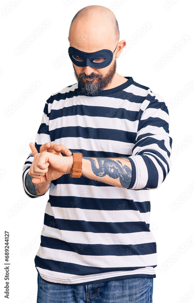 Poster Young handsome man wearing burglar mask checking the time on wrist watch, relaxed and confident