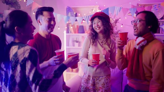 Friends Sit At Sofa Couch And Holding Drinks Joy Night Party With Pink Lighting At House Party.Young Asian Friends In Smart Casualwear Raising Arms While Dancing Together At Home Party In Living-room.