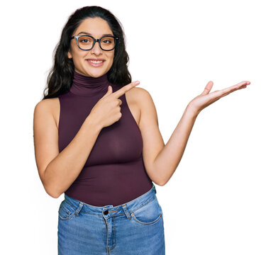 Brunette young woman wearing casual clothes and glasses amazed and smiling to the camera while presenting with hand and pointing with finger.