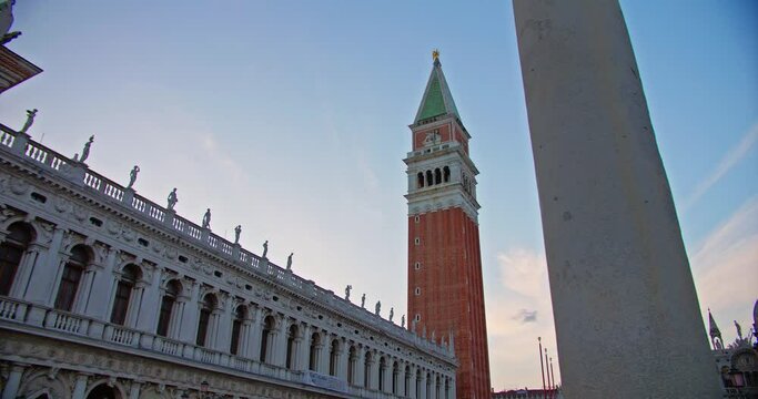 Dusk view of historical Procuratie Vecchie and the bell tower in St. Mark's Square, Venice. Statues atop ancient Venetian architecture with a clear sky.