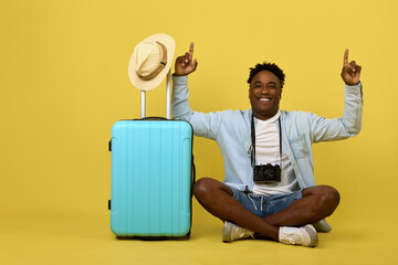 Young positive African photographer with a camera sits next to a travel suitcase. Cheerful stylish...