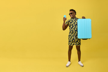 Happy handsome black man in glasses holding blue travel suitcase high in hand reading message on phone. On yellow background stands positive stylish African with mobile phone and suitcase in hand - obrazy, fototapety, plakaty