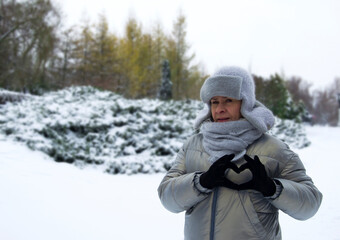 Fototapeta na wymiar Woman showing heart with hands. Valentine's day concept. 50 year old woman wearing earflap hat in winter park.