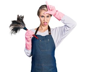Beautiful young blonde woman wearing apron holding cleaning duster making fun of people with fingers on forehead doing loser gesture mocking and insulting.