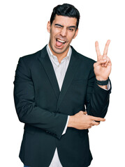 Handsome hispanic man wearing business clothes smiling with happy face winking at the camera doing victory sign with fingers. number two.