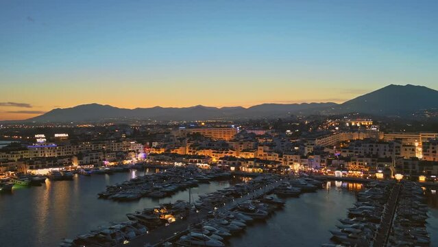 Establishing aerial panoramic view of Puerto Banus marina luxury port Marbella illuminated, Costa del Sol coastline Spain at blue hour. Travel vacation holidays in Spain