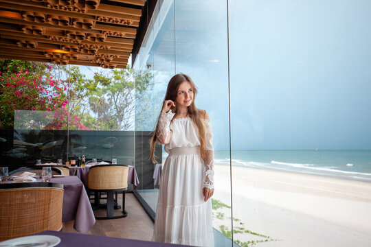 Serene beach scene with female in sophisticated attire standing inside upscale restaurant, reflecting on seaside tranquility. Luxury travel and lifestyle.