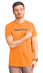 Handsome young man with bear wearing tshirt with happiness word message pointing with hand finger to the side showing advertisement, serious and calm face