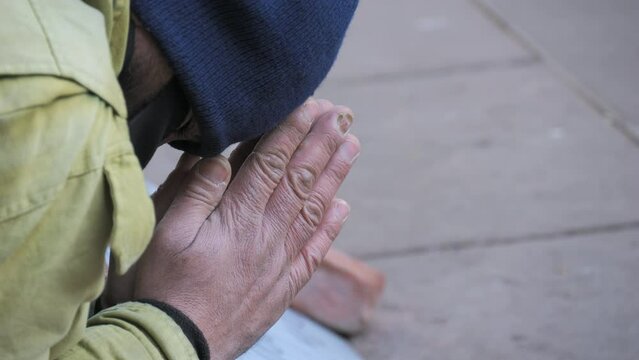 A poor homeless man begs for alms on his knees on the street in winter with his hands folded in a begging gesture. Crowds of people pass by slow motion
